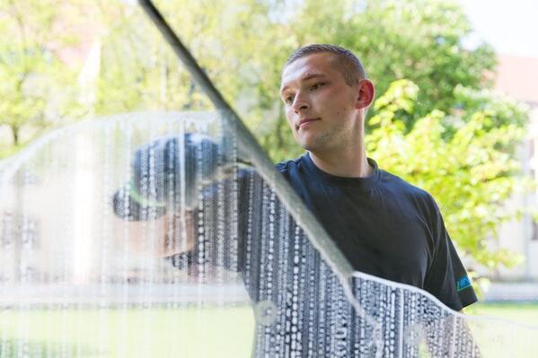 Glasreiniger säubert ein großes Fenster in einem modernen Gebäude. Das Fenster spiegelt den grünen Außenbereich wider.