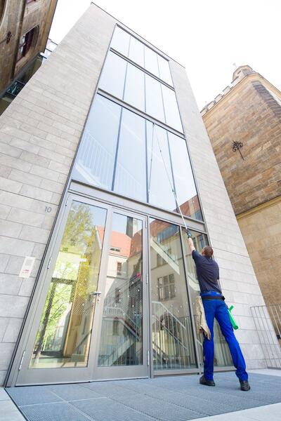 Mann in Arbeitskleidung reinigt mit einer Teleskopstange die Glasfassade eines hohen, modernen Gebäudes.