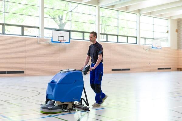 Mann in blauer Arbeitskleidung bedient eine blaue Wischmaschine auf dem Boden einer Turnhalle mit Sportlinien.